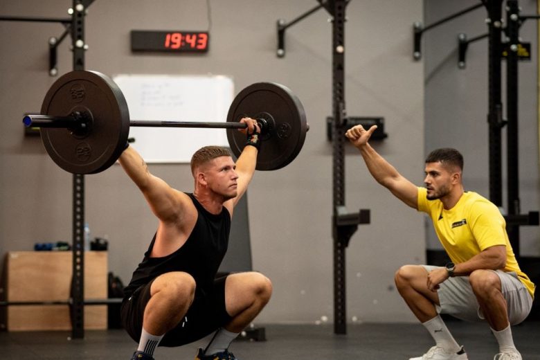 Athlete performing weightlifting exercise under trainer's guidance in a gym setting.